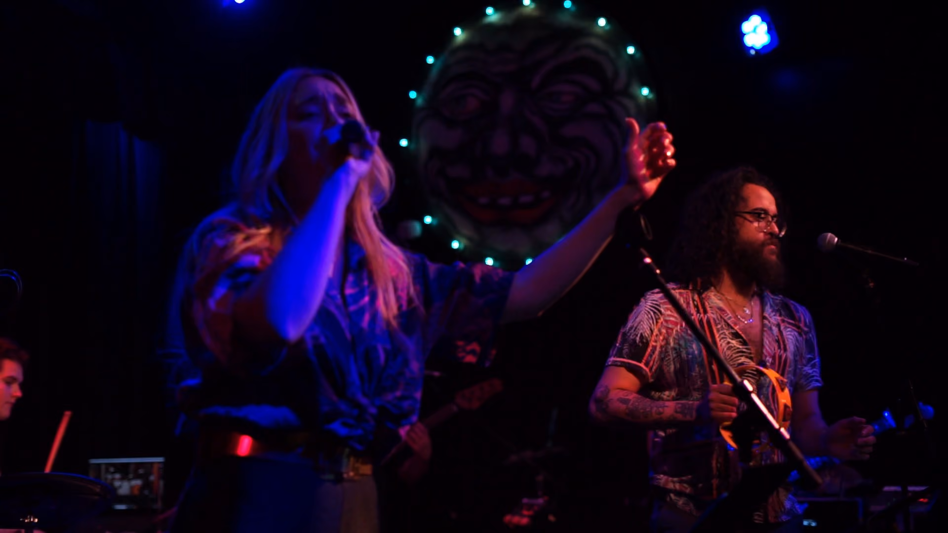 The band on stage under blue and pink lights, a ring of bulbs glowing behind the drums